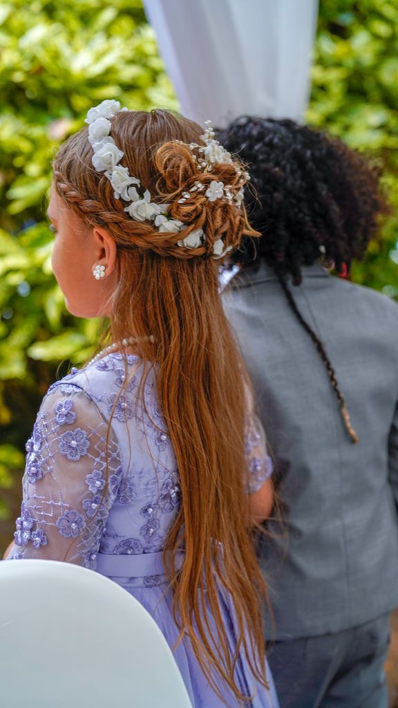 Young girl in a purple dress with floral hair at an outdoor wedding ceremony.
